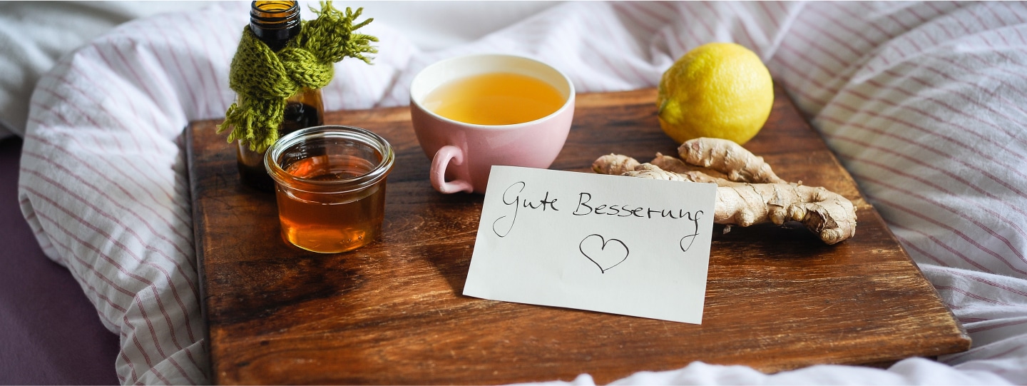 Holztablett auf Bett: mit Honig, Tee, Zitrone, Ingwer, kleine Flasche, Zettel “Gute Besserung”